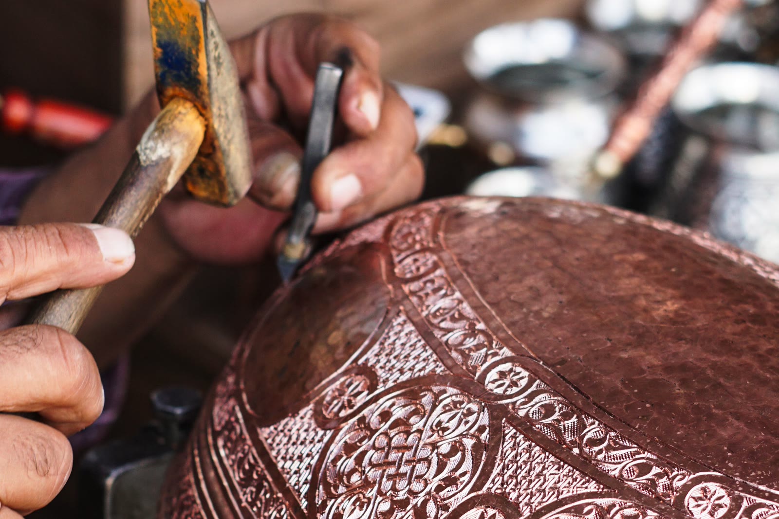 Two hands holding a chisel and hammer, chasing a fine repeating pattern into a copper tray