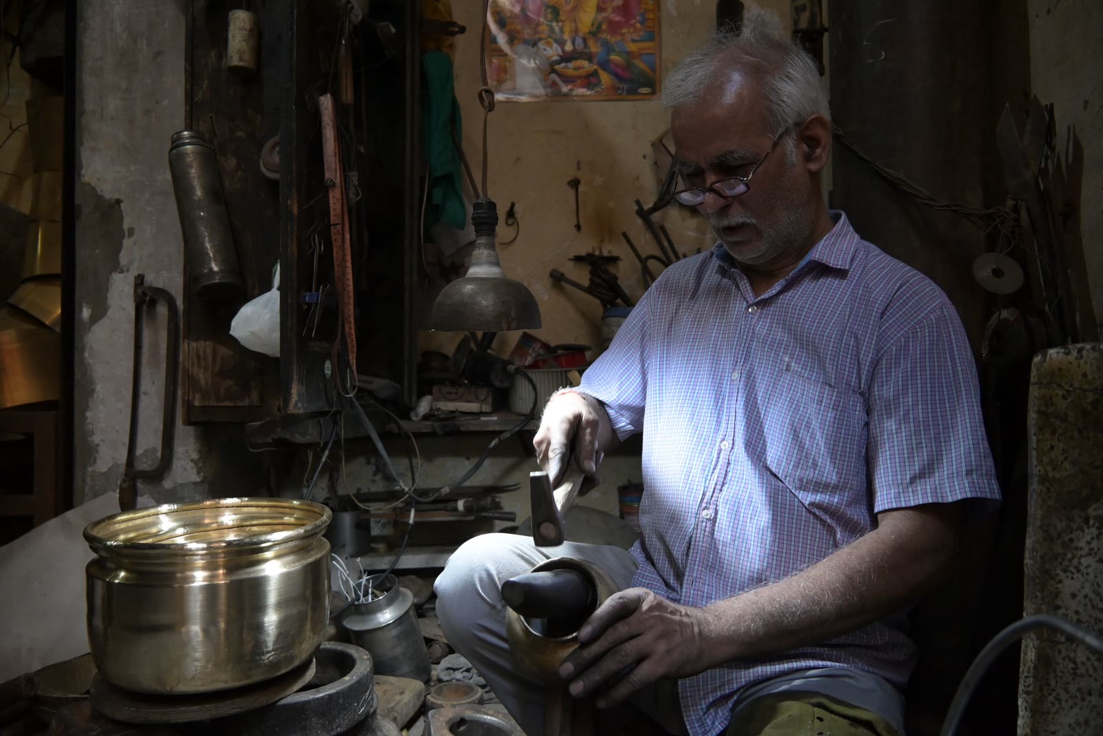 A metalworker at his bench in a brass workshop, a finished pot beside him and tools racked on the wall